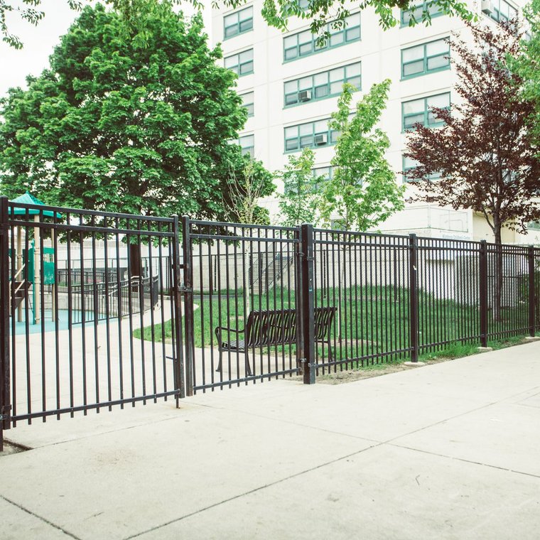Gated entry to playground with metal benches, lush green grass, trees and plastic play gym